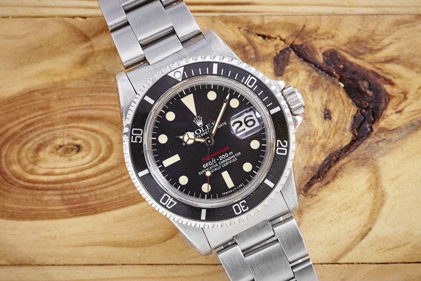 A vintage Rolex "Red" Submariner dive watch with a black bezel and dial, white markers, and a date magnifier, showing ROLEX branding and set to the 24th, displayed on a wooden surface.
