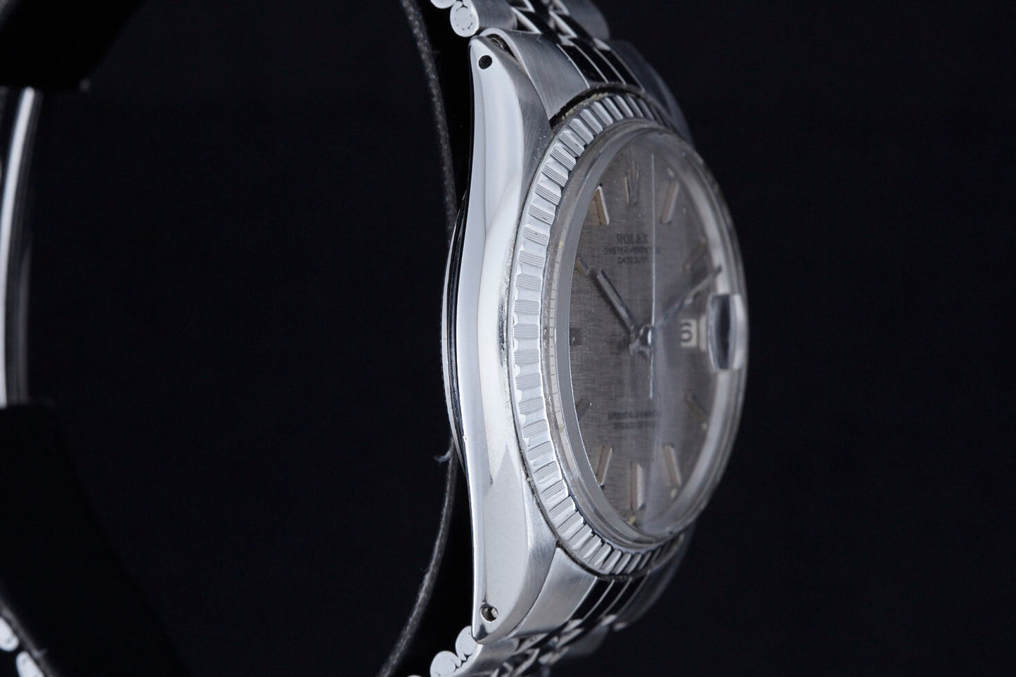 Close-up side view of the Rolex Datejust 1601 Grey Linen by Rolex, featuring a silver metallic band, textured bezel, and grey linen watch face with time and date display against a black background.