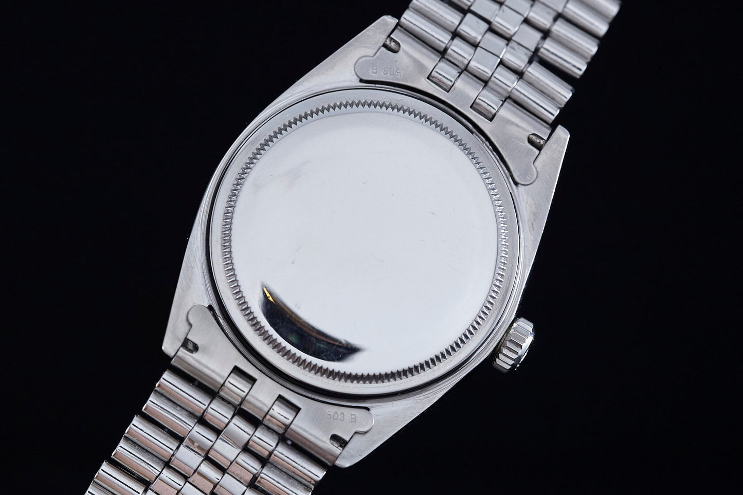 A close-up of the back of a Rolex Datejust 1601 Grey Linen highlights its smooth, unmarked stainless steel case back and crown with a metal link band, showcasing classic Rolex design against a dark background.