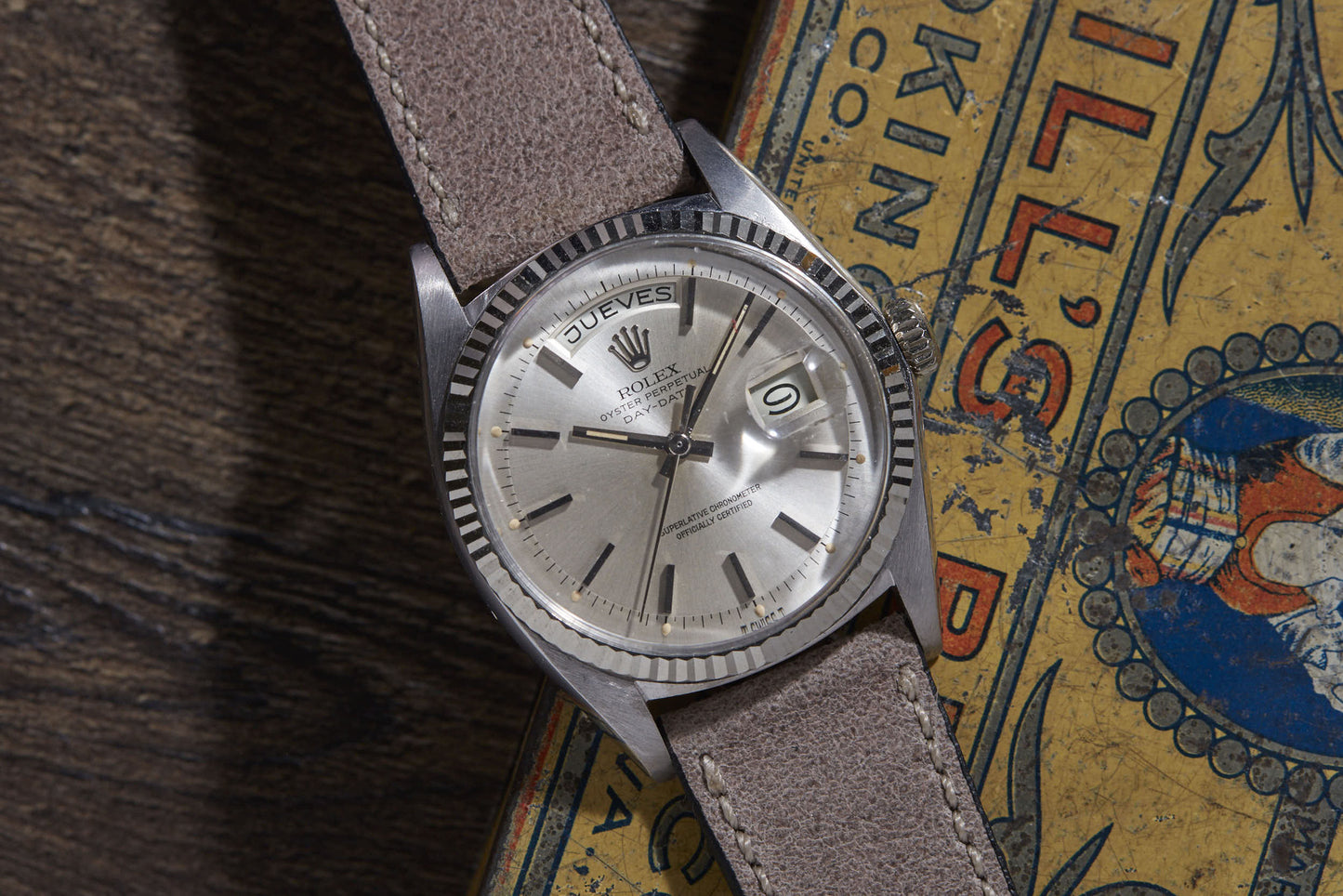 A Rolex Day Date wristwatch by Rolex with automatic movement and a light brown leather strap sits on a colorful vintage book. The Reference 6511 shows the day in Spanish (JUEVES) and the date (9).