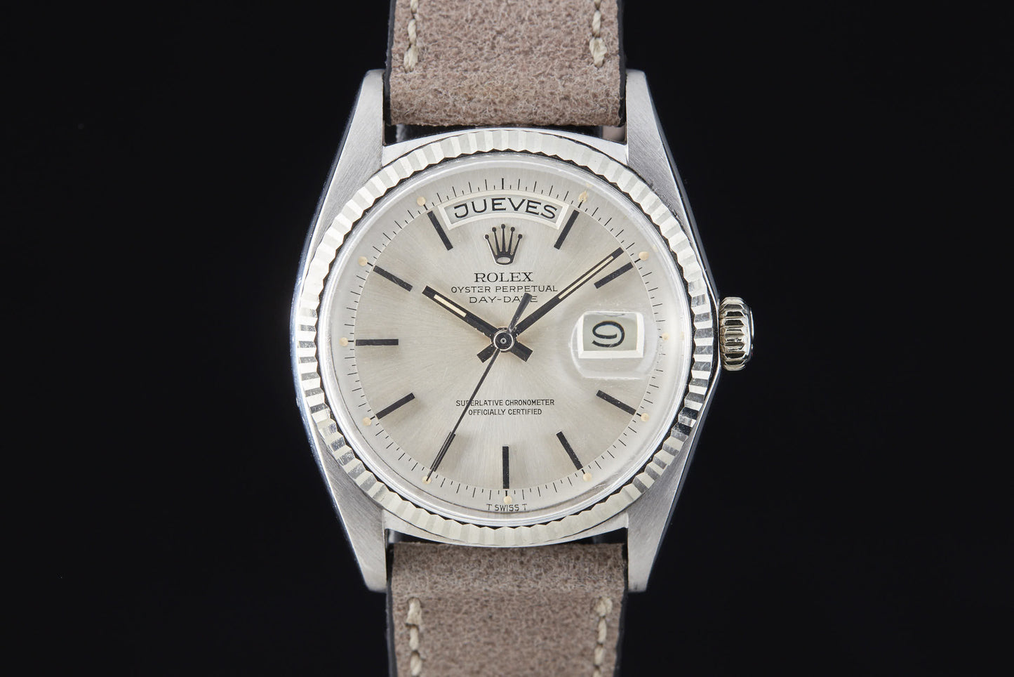 The Rolex Day Date by Rolex features a silver case, beige dial, Spanish day display ("JUEVES"), magnified date at 3 o’clock, brown leather strap, silver hour markers, automatic movement (Ref. 6511), shown on a black background.