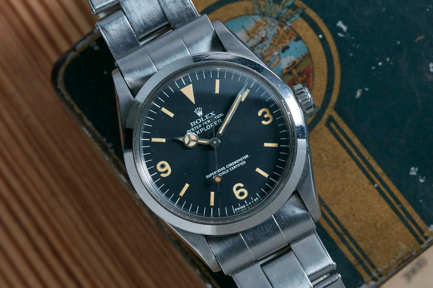 The Rolex Explorer by Rolex featuring a black 3-6-9 Arabic dial, luminescent hour markers, and stainless steel band is displayed on a weathered decorative box.