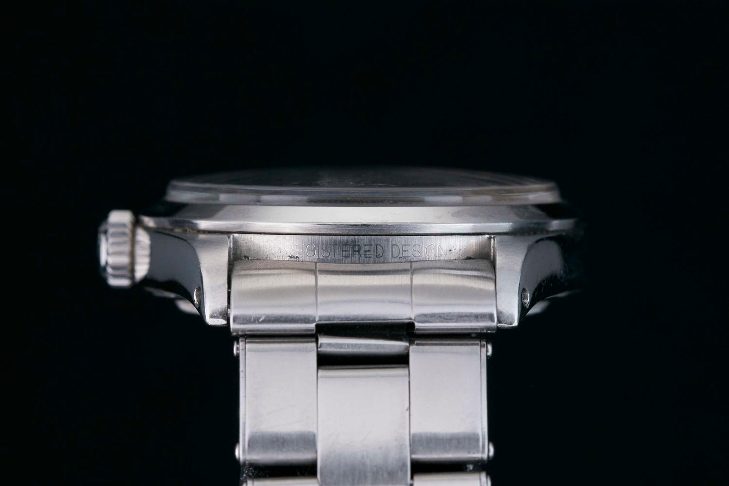 Close-up side view of a silver Rolex Explorer wristwatch with a metal band, displaying the case, crown, and bracelet against a black background. Faint engraved text appears on the case with a classic black 3-6-9 Arabic dial. Brand: Rolex.