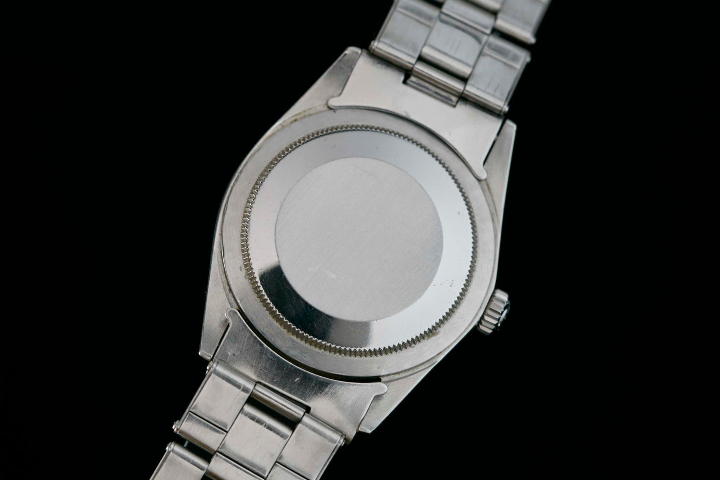 A close-up of the back of a stainless steel Rolex Explorer wristwatch with a metal link band, set against a black background. The plain, unmarked caseback highlights the signature quality of the Rolex brand.