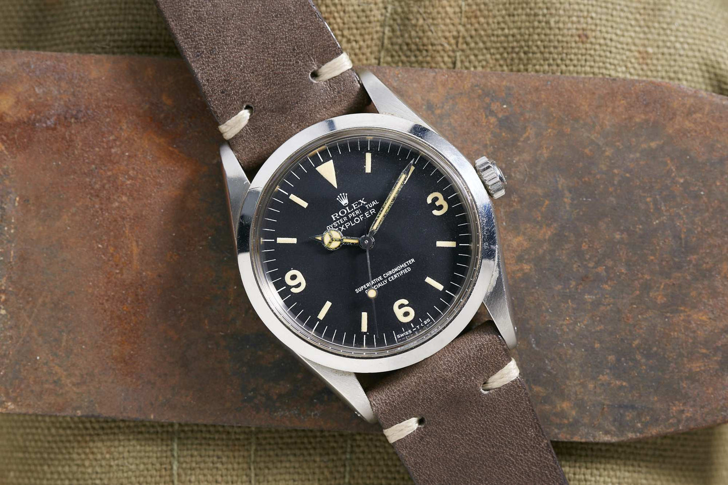 A Rolex Explorer 1016 with a black dial, silver case, and brown leather strap rests on rusted metal over textured fabric. Detailed Description Coming Soon!.