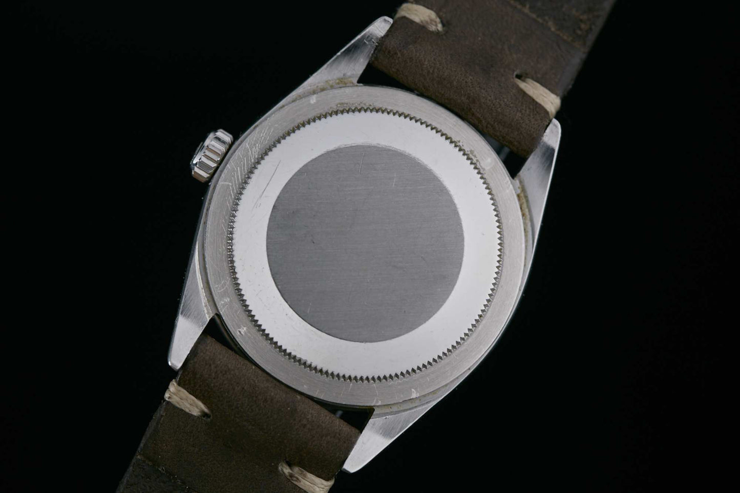 A close-up of the Rolex Explorer 1016 by Rolex, featuring a plain, round metal case back with a ridged edge and a brown leather strap, set against a black background. Detailed Description Coming Soon!.