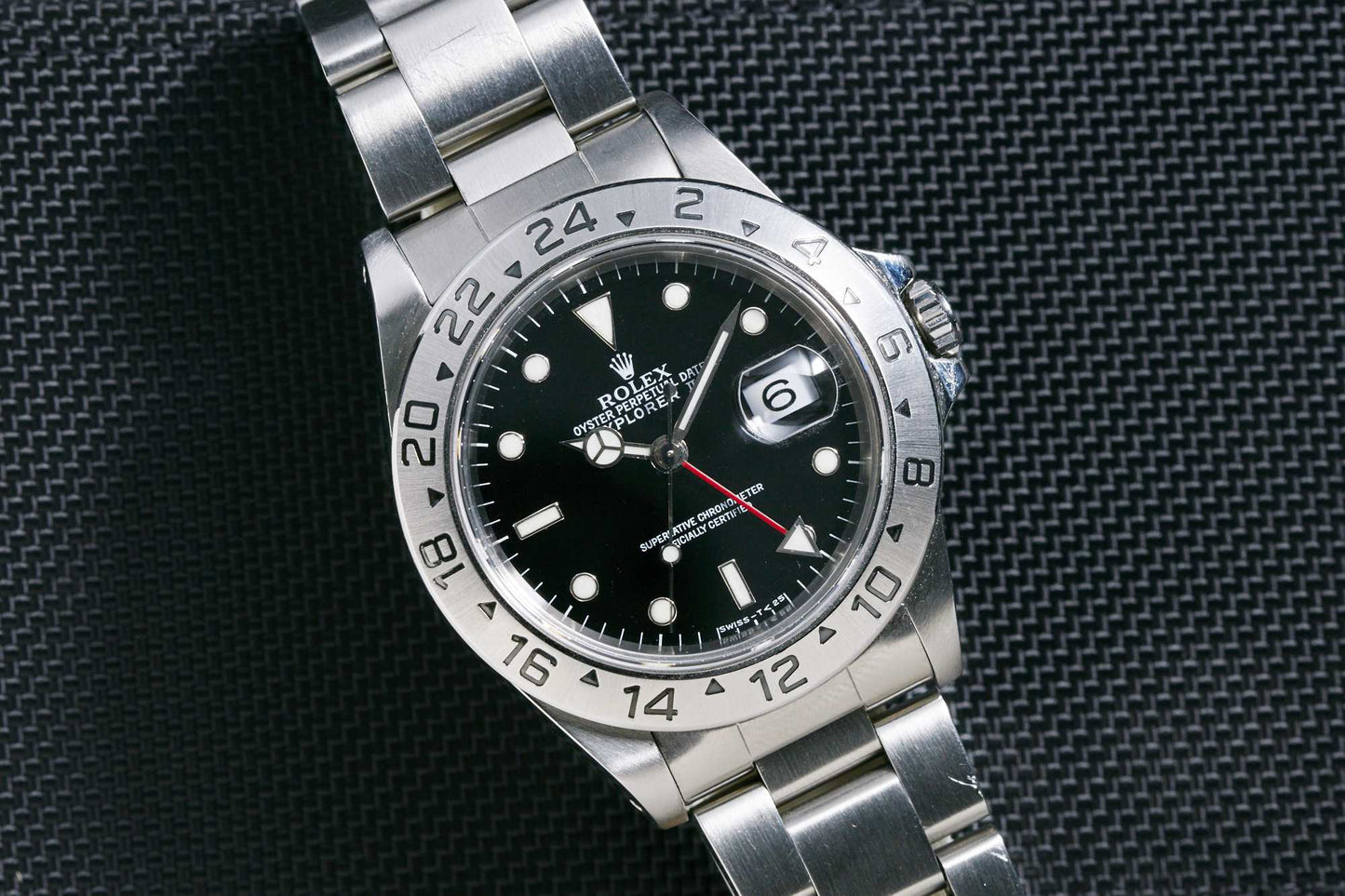 The Rolex Explorer II by Rolex features a stainless steel case, black dial, silver hour markers, date window at 3 o’clock, 24-hour bezel, and metal link bracelet. It's displayed on a textured dark surface.