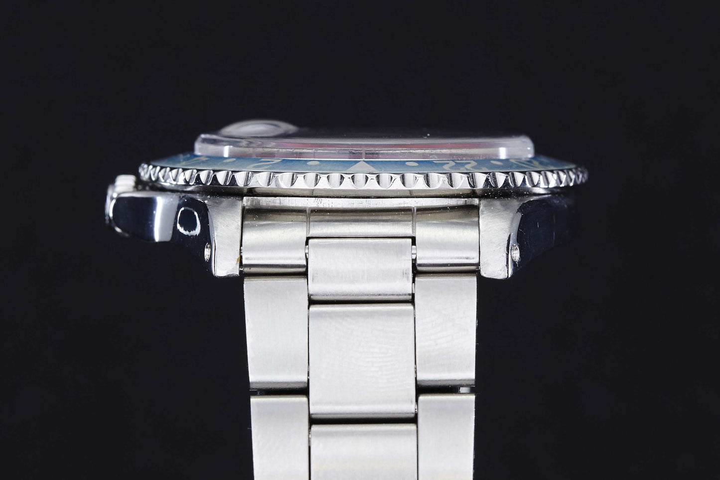A close-up side view of the Rolex GMT Master 1675 by Rolex, featuring its stainless steel case, serrated bezel, and crown on a metal bracelet against a black background.