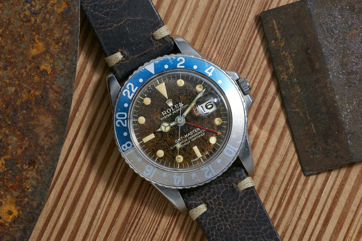 A Rolex GMT-Master sport watch by Rolex, featuring a blue and white bezel, cracked leather strap, and worn brown dial, sits on a wooden surface next to two rusty metal plates.
