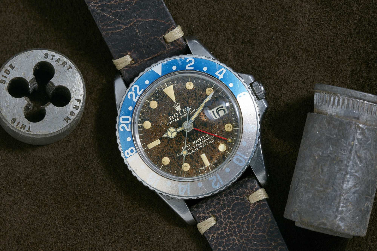A Rolex GMT-Master wristwatch with faded blue and white bezel, aged brown dial, and leather strap sits on a brown surface next to a metal tool, highlighting the classic style of this iconic Rolex sport watch.
