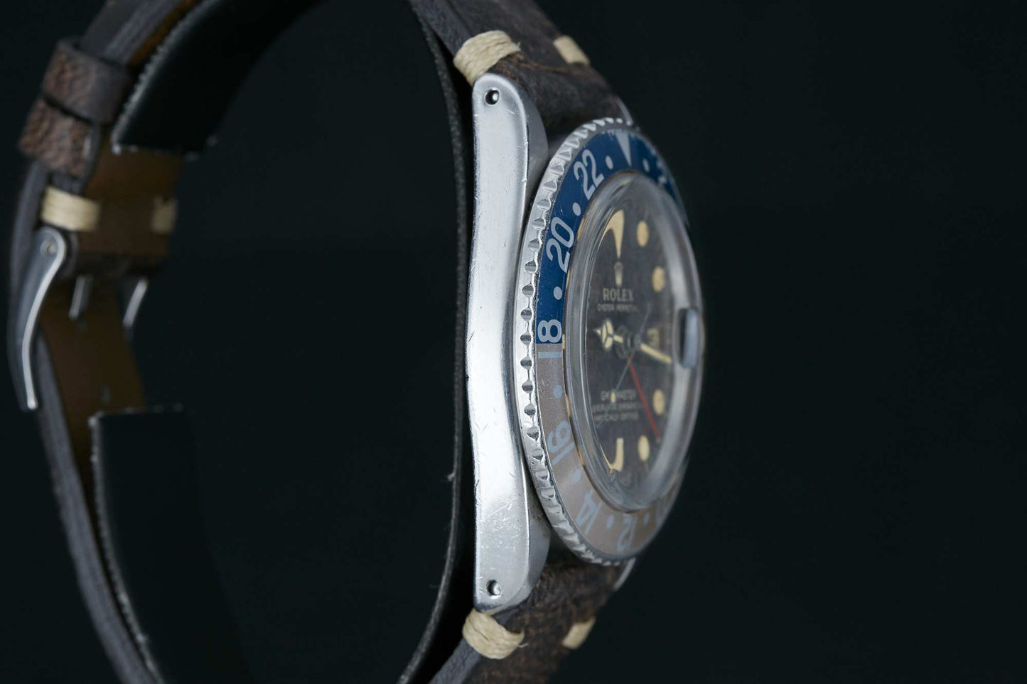 A close-up side view of the Rolex GMT-Master by Rolex, featuring a blue and silver bezel, brown leather strap, and worn watch face and bezel details against a black background.