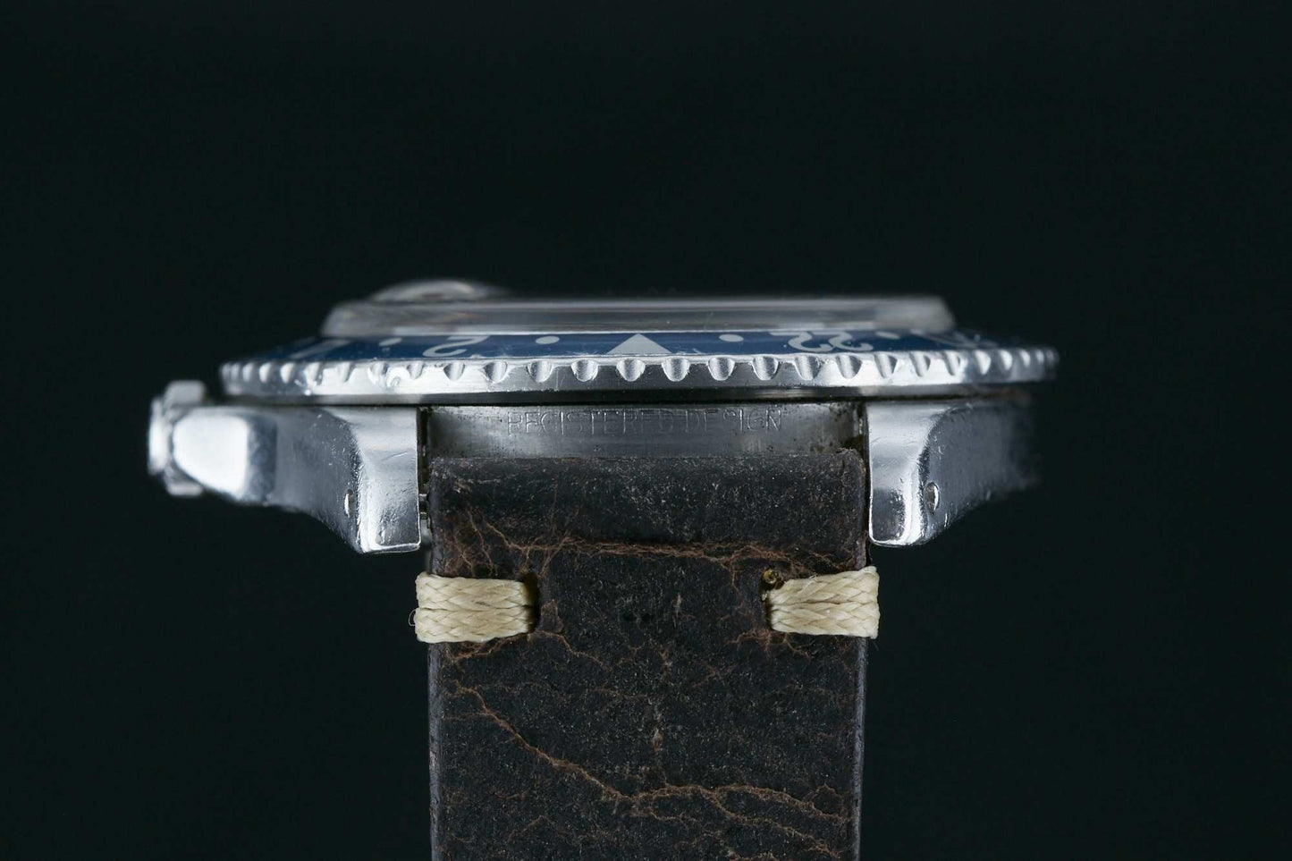 Side view of a vintage Rolex GMT-Master wristwatch with a weathered dark brown leather strap stitched in light thread, featuring a textured bezel and metal case against a black background. Ideal for Rolex sport watch enthusiasts.