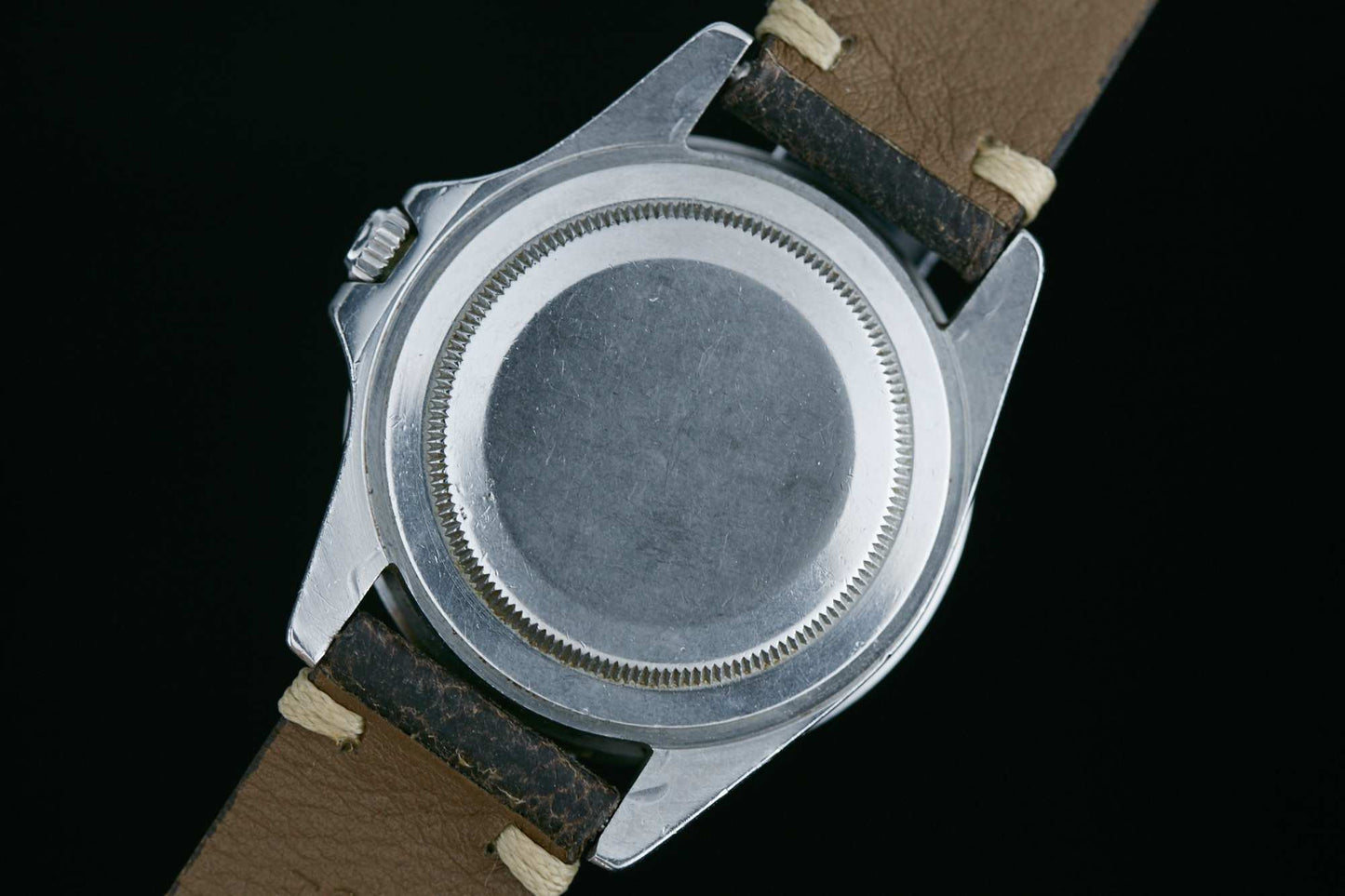 The image shows the back of a silver Rolex GMT-Master wristwatch with a round, smooth case and tan leather strap, set against a black background. Its classic design captures the understated elegance of vintage Rolex sport watches.