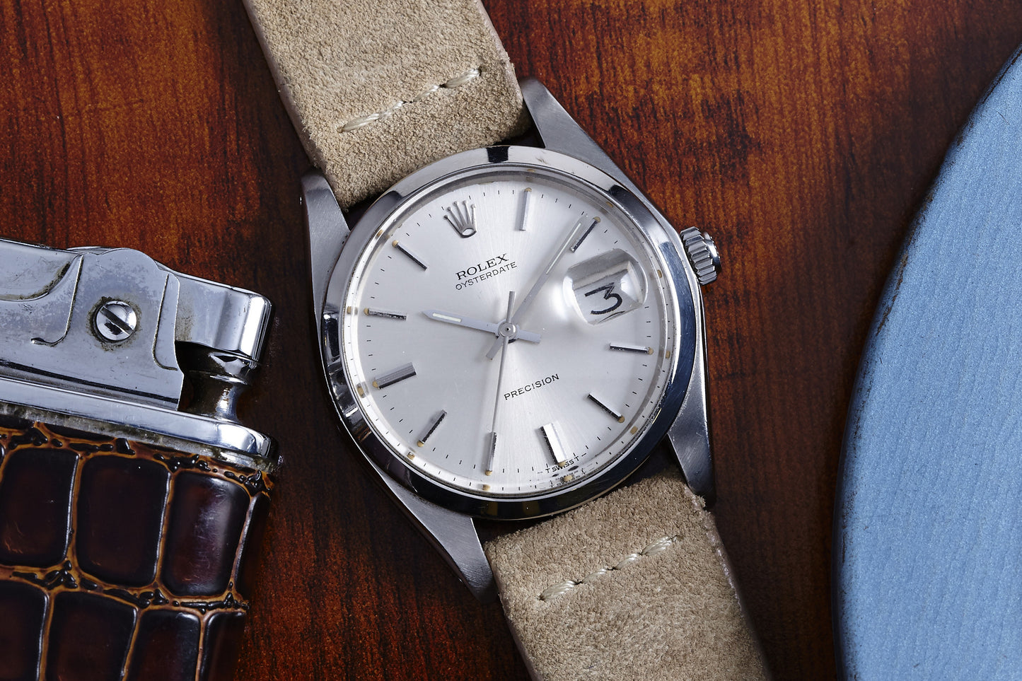 A 1978 Rolex Oysterdate Precision with a beige suede strap sits on a wooden surface beside a metal lighter with a brown textured case and the edge of a blue circular object. The watch displays the date and reads 3:10.