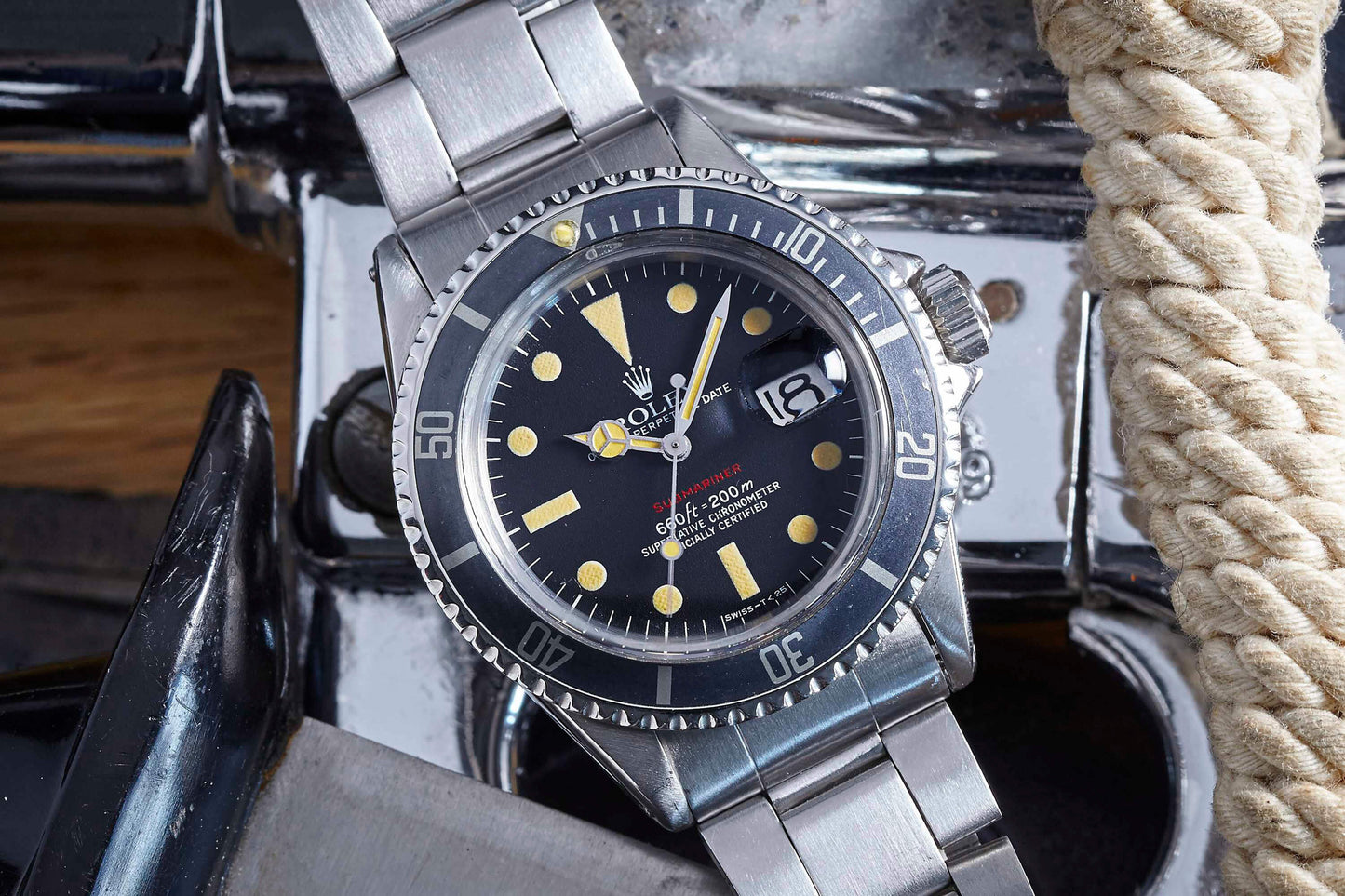 A close-up of the Rolex Submariner by Rolex features its stainless steel bracelet, black dial, and rotating bezel as it rests on a shiny metallic surface beside coiled rope.