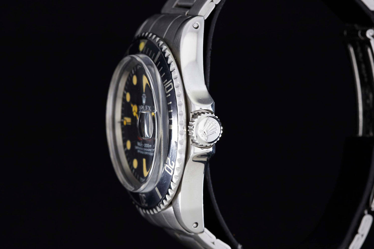 A close-up side view of the Rolex Submariner by Rolex shows its luxury dive watch design, featuring a silver metal case, black bezel, visible crown, yellow hour markers, and white hands against a black background.
