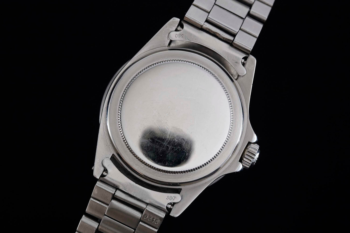 A close-up of the back of a Rolex Submariner dive watch by Rolex, featuring a stainless steel caseback, metal bracelet, and right-side crown against a black background.