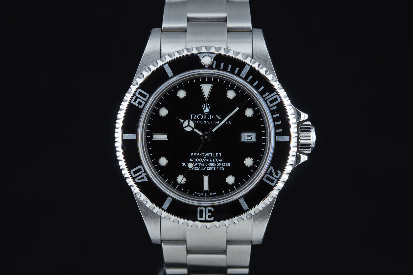 The Rolex Sea Dweller by Rolex is designed for deep diving, featuring a black dial with white markers and hands, a date window at 3 o’clock, a silver bezel, and a metal bracelet band—originally inspired by COMEX divers.