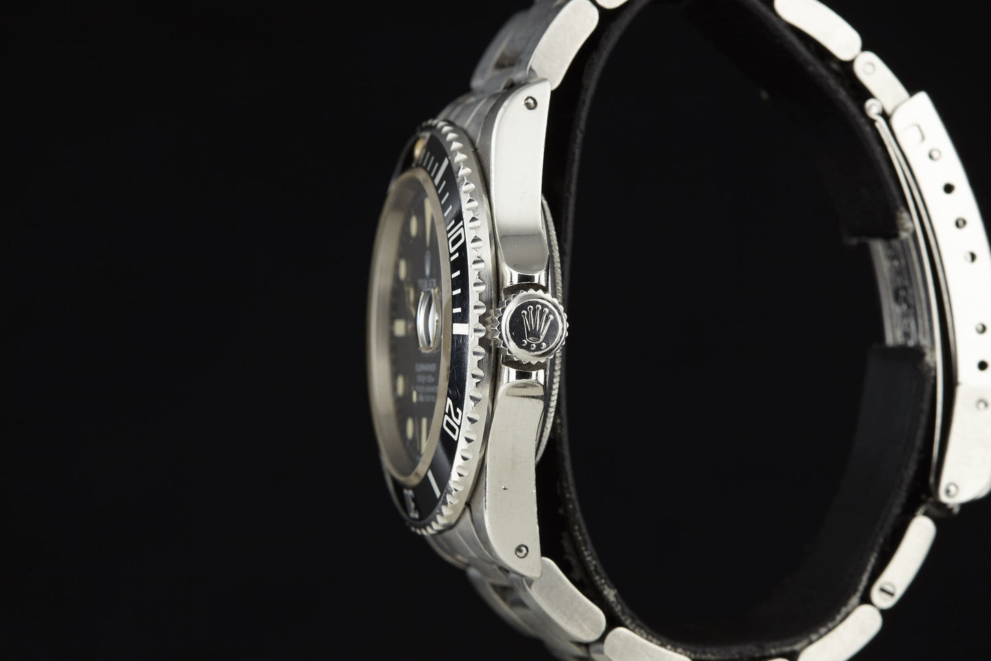 A close-up side view of the Rolex Submariner luxury watch with a black bezel and metal strap, featuring the Rolex crown logo on the winding crown, set against a black background.