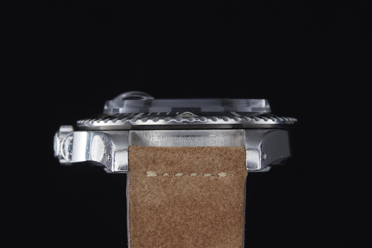 Side view of the Rolex Submariner dive watch by Rolex, featuring a metallic case, gear-edged bezel, and brown suede leather strap against a black background. The crystal, crown, and engraved case-side text are visible.