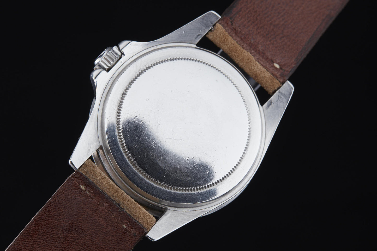 A close-up of the back of a Rolex Submariner by Rolex, featuring a silver case and brown leather strap, displays its smooth, round caseback against a black background.