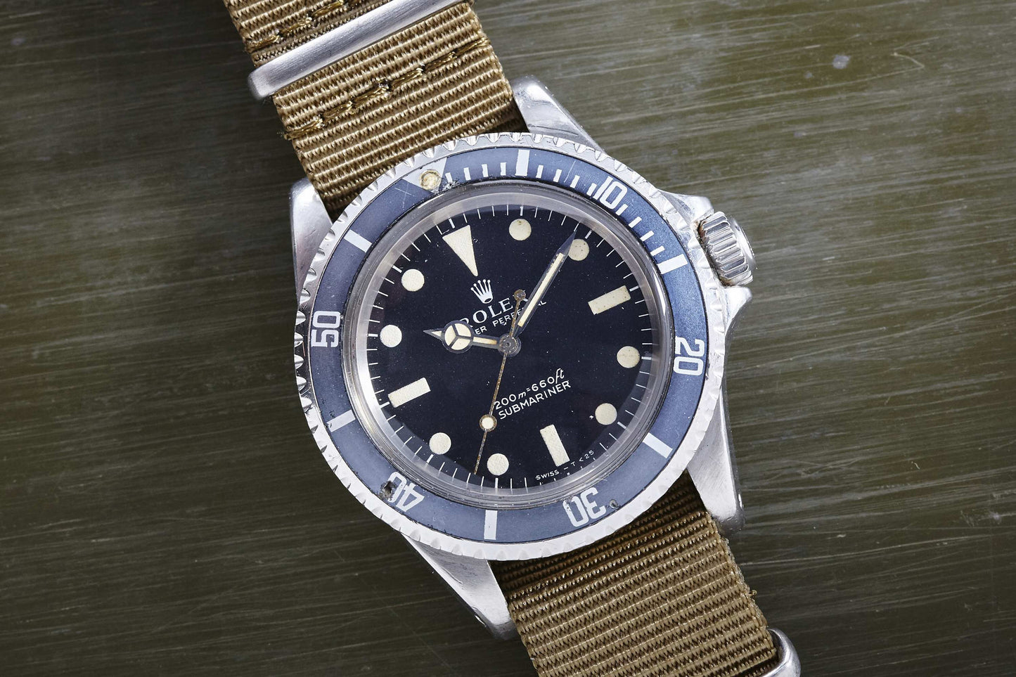 A Rolex Submariner watch by Rolex, featuring a black dial, round hour markers, rotating bezel, and khaki NATO strap, is shown on a textured green surface—a timeless tool watch for classic style enthusiasts.