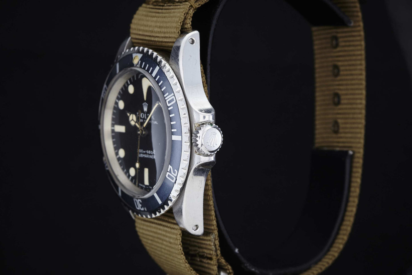 A close-up side view of the Rolex Submariner by Rolex, a silver tool watch with a black bezel and round face, featuring a khaki fabric strap, set against a black background.