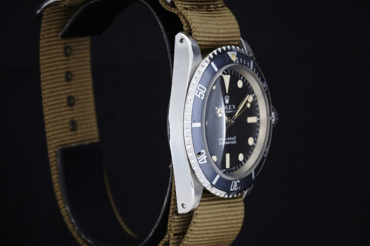 Close-up side view of the Rolex Submariner by Rolex, featuring a black dial, rotating bezel, and a brown NATO-style fabric strap against a black background.