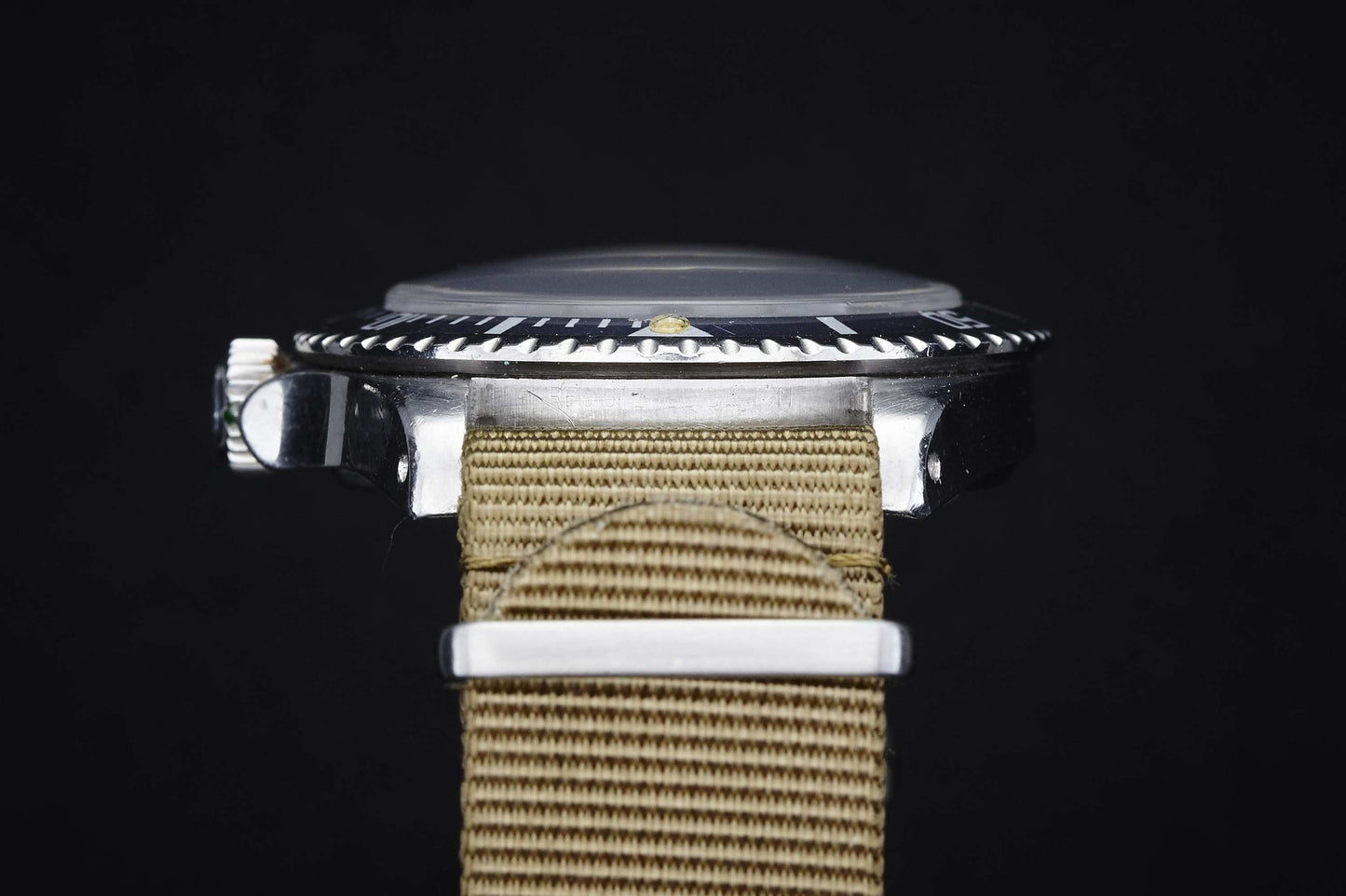 Close-up side view of the Rolex Submariner by Rolex, featuring a silver metal case, ridged bezel, domed glass crystal, and beige woven fabric strap set against a solid black background.