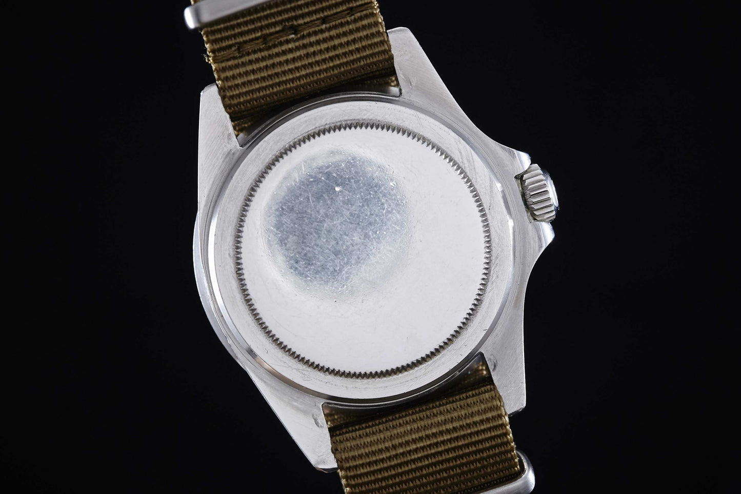 The image shows the back of a Rolex Submariner, a stainless steel tool watch by Rolex, featuring a round, unmarked case and a brown fabric strap, set against a black background.