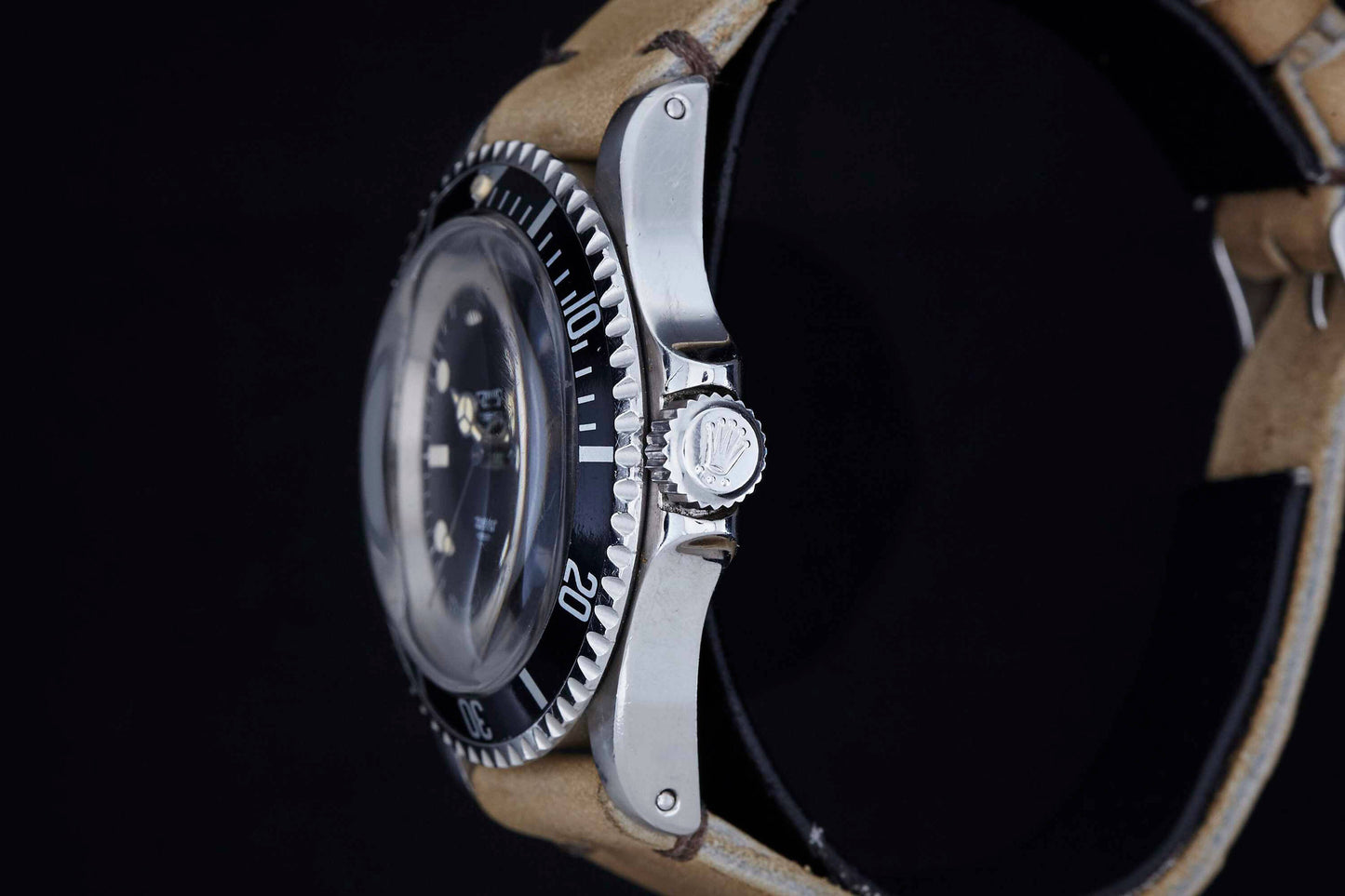 Side view of a Rolex Submariner wristwatch by Rolex, featuring a black bezel, clear glass face, beige leather strap, and crown with the Rolex logo, all set against a black background.