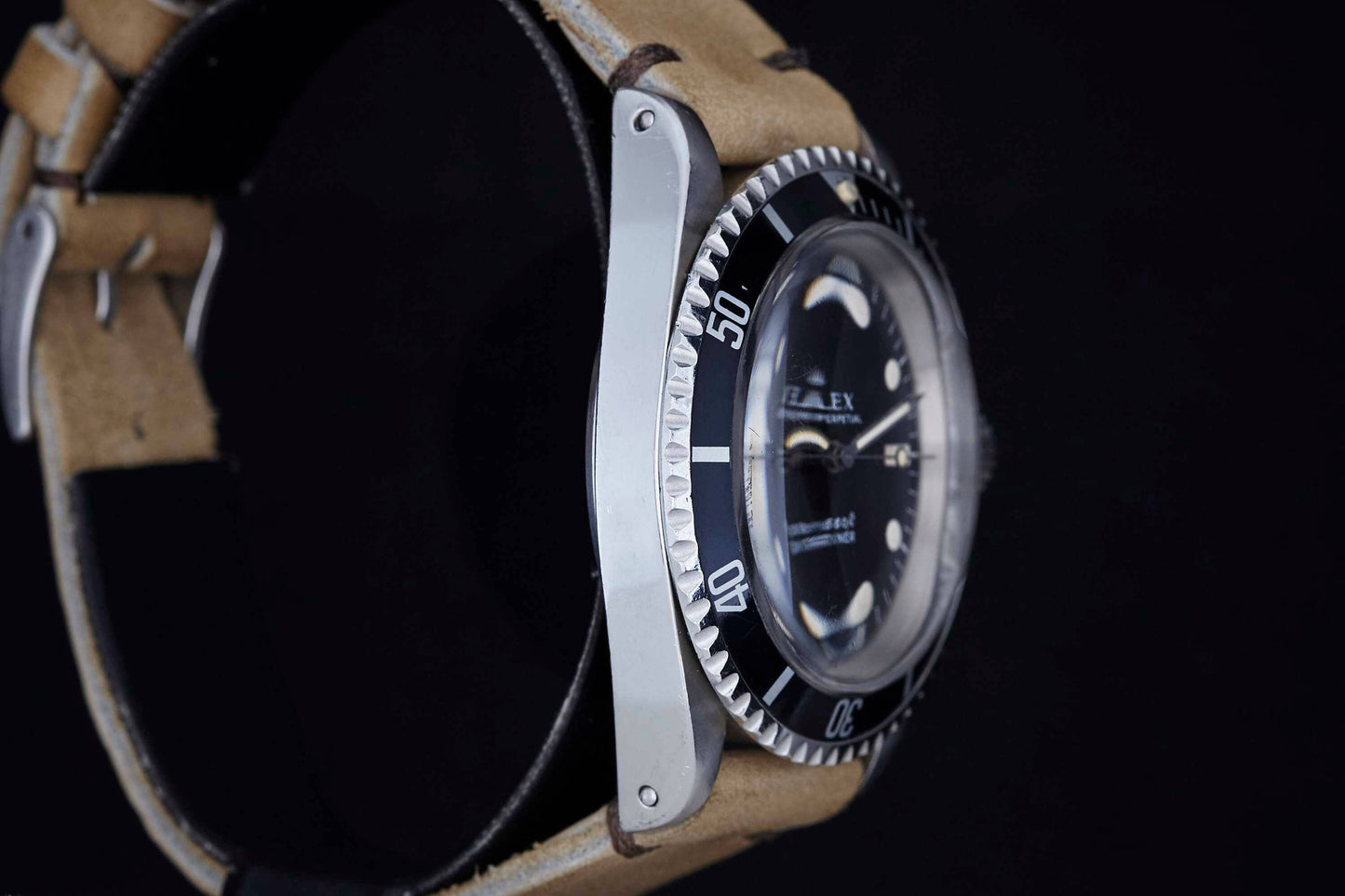 A close-up side view of the Rolex Submariner by Rolex, featuring a black bezel, silver case, and tan leather strap against a black background. The watch face is partly visible, showing minute markings and select hour markers.