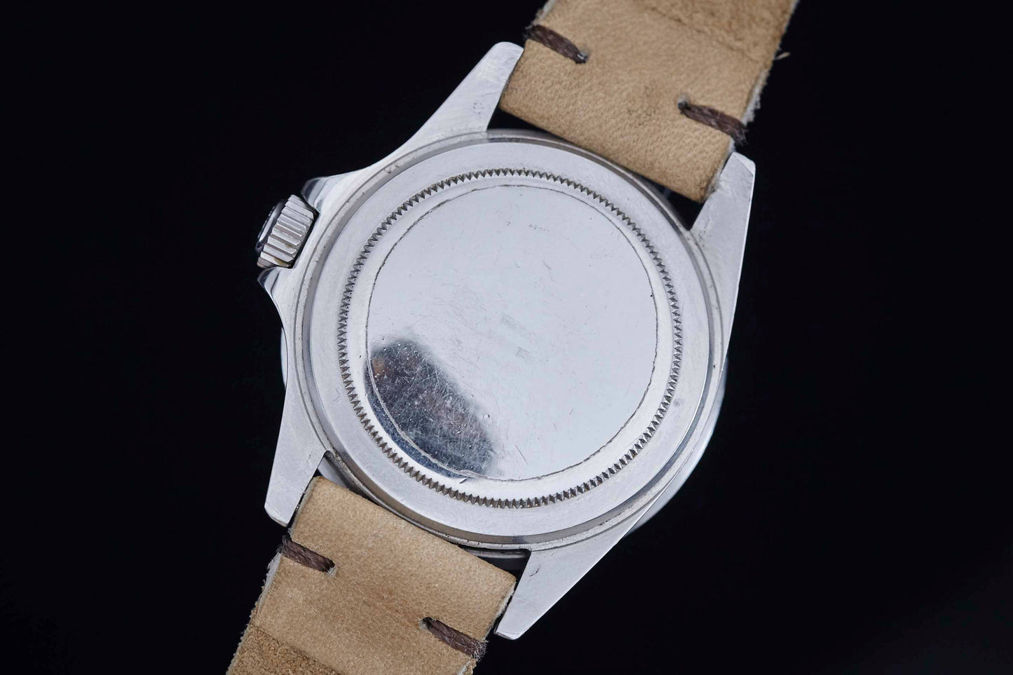 A close-up of the back of a Rolex Submariner by Rolex shows its round, plain metal case and light tan leather straps against a black background. The slightly scratched, unmarked case hints at this timepiece’s storied past.