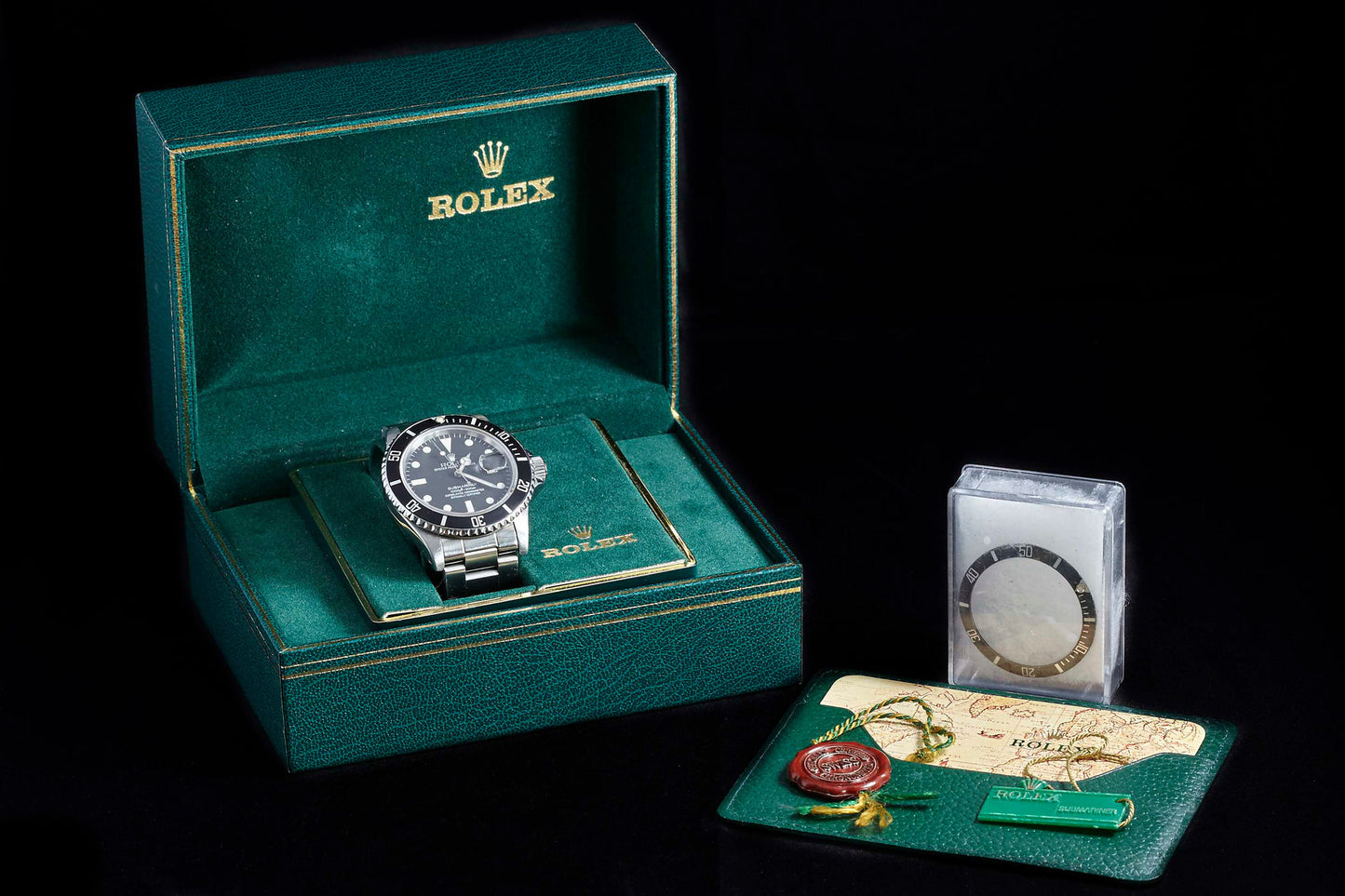 A Rolex Submariner Date by Rolex in a green box with gold trim, includes a separate bezel, red wax seal, green tag, and guarantee card, all displayed against a black background.