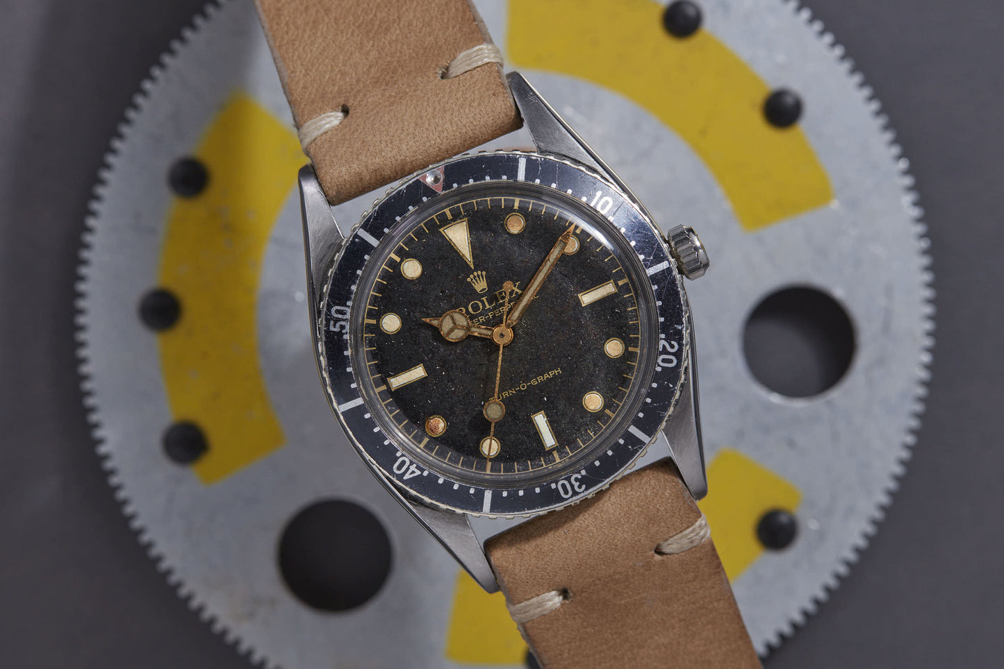 A Rolex Turn-O-Graph wristwatch by Rolex, featuring a black dial, gold hour markers, and a tan leather strap, sits atop a round metallic object with yellow and black segments.