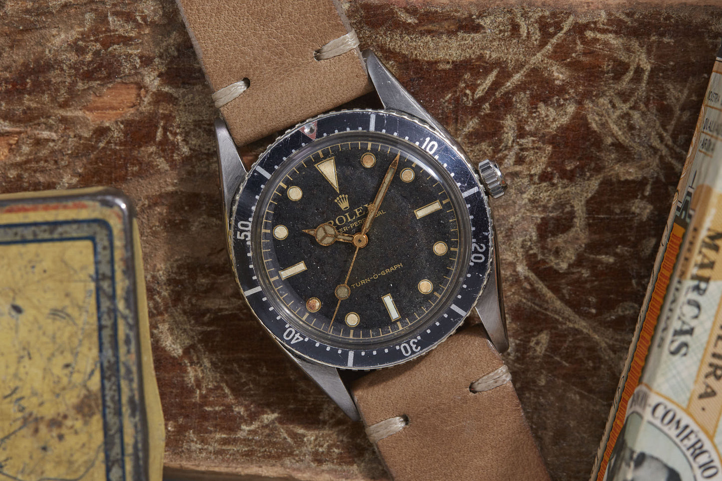 A Rolex Turn-O-Graph wristwatch by Rolex, featuring a black dial, tan markers, brown leather strap, and minute-marked rotating bezel, rests on a worn wooden surface among vintage items.