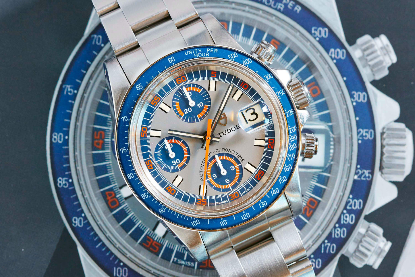 A Tudor Monte Carlo 9420/0 chronograph watch with a blue and orange bezel, silver dial, and metal bracelet rests atop a similar watch image, highlighting the refined vintage details of Tudor craftsmanship.