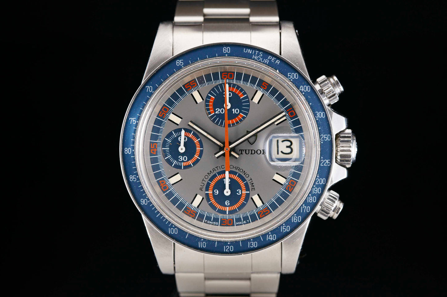 A close-up of the Tudor Monte Carlo 9420/0 chronograph shows its stainless steel case, blue and orange-accented dial with three subdials, a date window at 3 o'clock, and a blue tachymeter bezel—reflecting Tudor’s Rolex-quality craftsmanship.