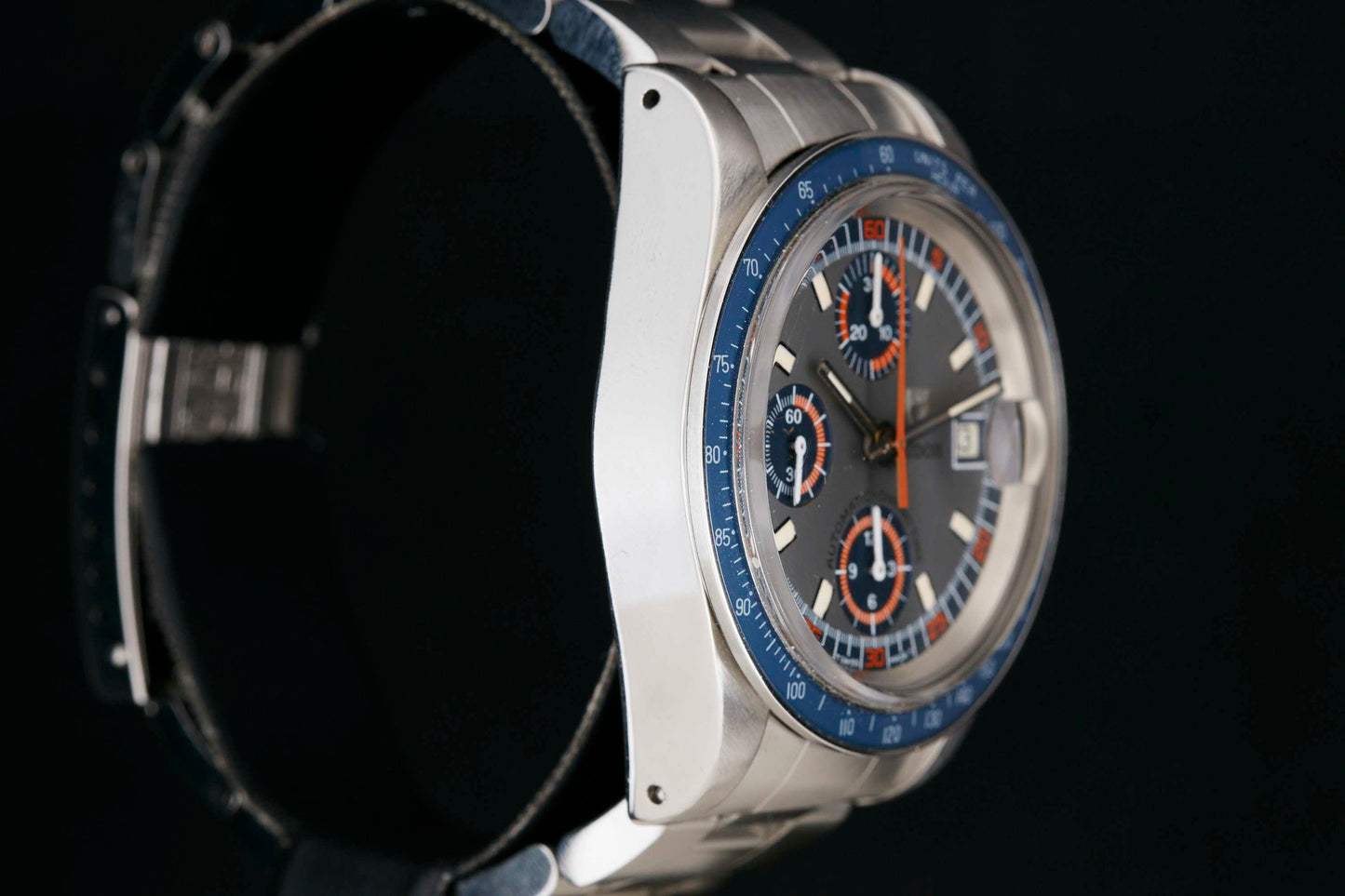 A close-up of the Tudor Monte Carlo 9420/0 highlights its silver case, blue and orange bezel, black and orange subdials, date window, and stainless steel band—showcasing Tudor’s vintage style and renowned craftsmanship against a black background.