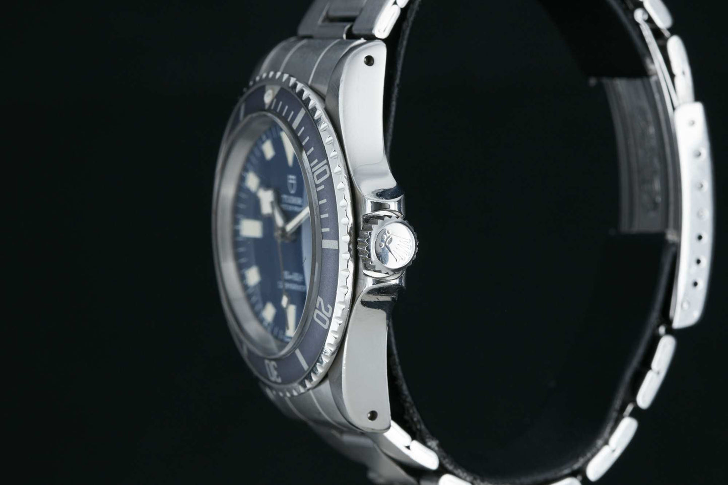 A close-up side view of the Tudor Submariner Snowflake by Tudor, featuring a blue dial, stainless steel case and bracelet, with crown and bezel details visible against a black background. Detailed Description Coming Soon!.