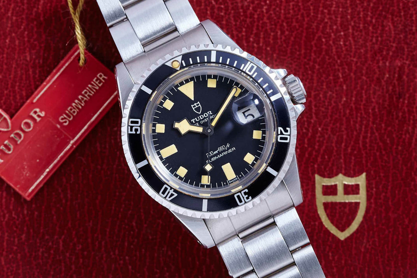 A Tudor Snowflake Submariner by Tudor, with a stainless steel band, black dial, yellow hour markers, and date window, rests on a red textured background beside a Tudor tag and shield, highlighting its classic Oyster case design.