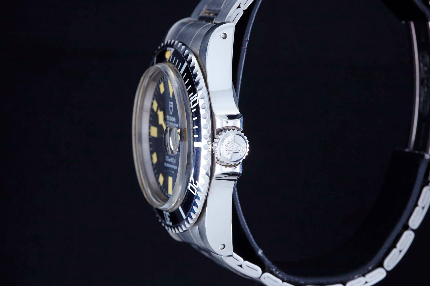A close-up side view of the Tudor Snowflake Submariner shows its silver case, black and yellow dial, metal bezel, and stainless steel bracelet against a black background, highlighting the engraved Tudor logo on the crown.