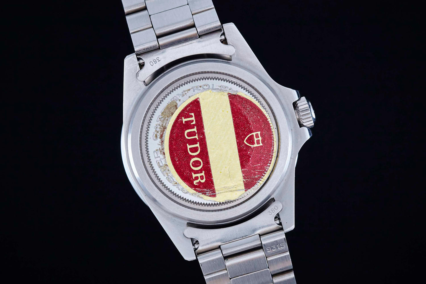 Close-up of the back of a Tudor Snowflake Submariner, featuring a caseback sticker with the Tudor logo in red and yellow on a stainless steel bracelet, inspired by the Rolex Oyster case, set against a black background.