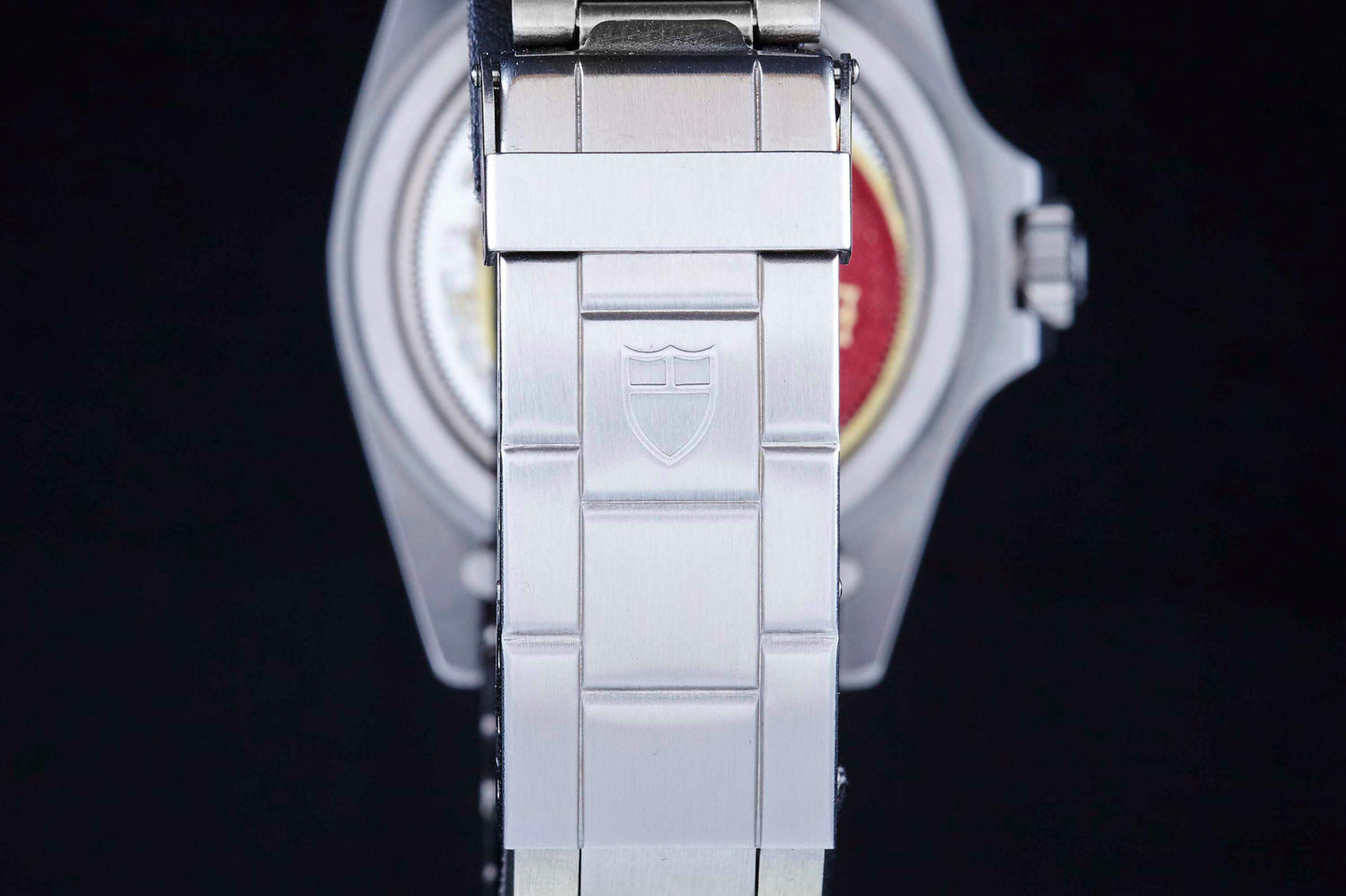 A close-up of a silver metal Tudor bracelet with the shield logo engraved on its clasp, set against a dark background. The Tudor Snowflake Submariner case and crown are partially visible in the background.