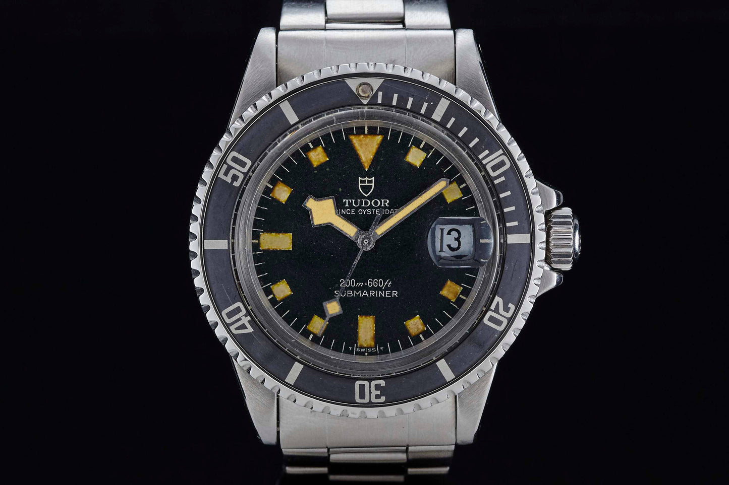 A close-up of the Tudor Submariner by Tudor, showing its black dial, prominent yellow hour markers, date window at 3 o'clock, and a stainless steel case and bracelet set against a black background.
