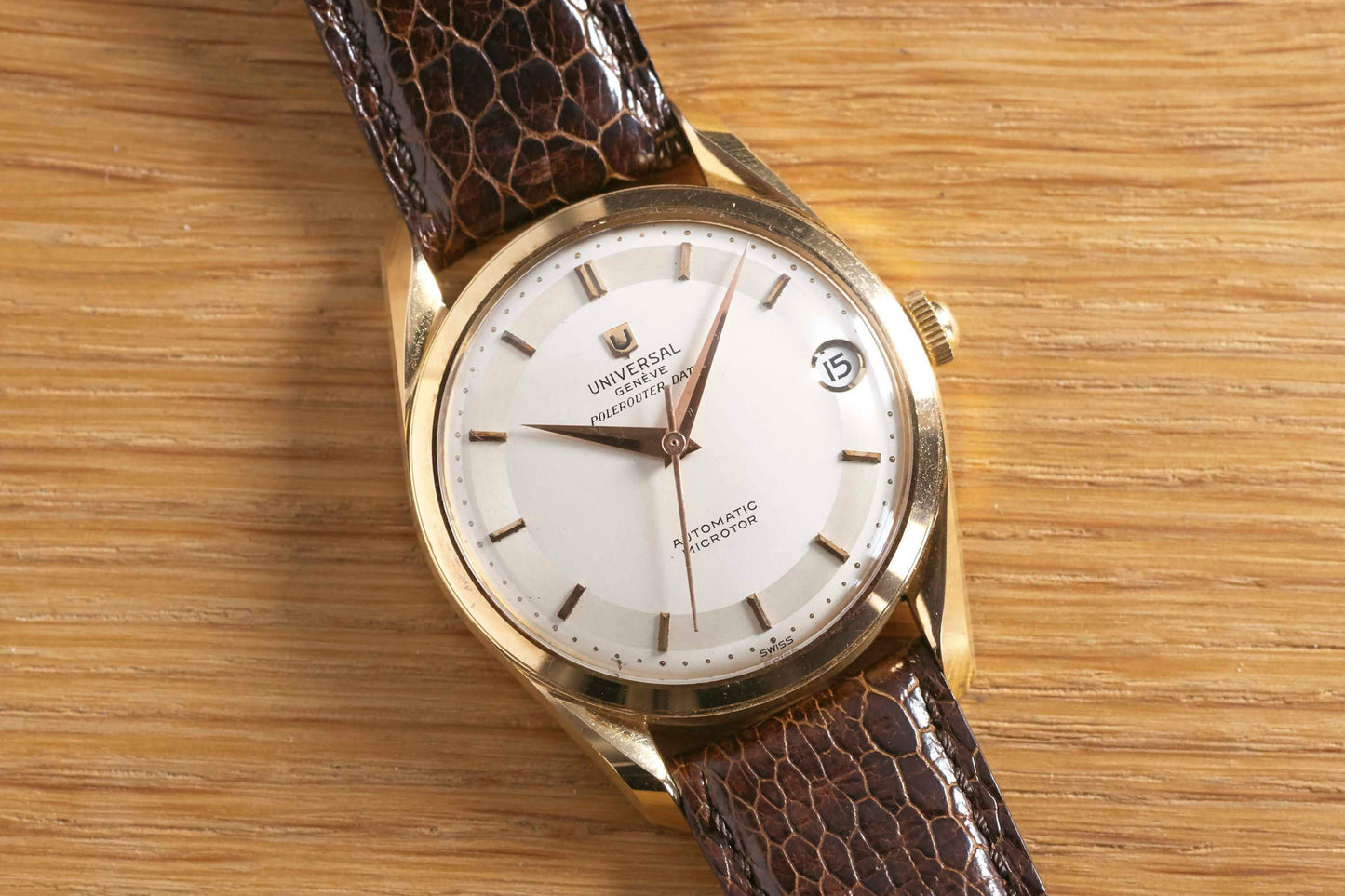 The Universal Genève Polerouter Date by Universal Geneve features a gold case, white dial, brown leather strap, and date window at 3 o’clock—a classic vintage dress watch shown elegantly on a wooden surface.
