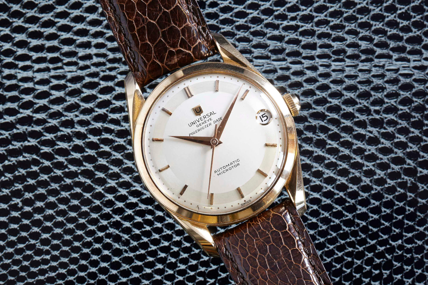 The Universal Genève Polerouter Date, a vintage dress watch by Universal Geneve, features a gold case, white dial with gold hour markers, date window at 3 o’clock, and brown leather strap, shown on a textured dark surface.