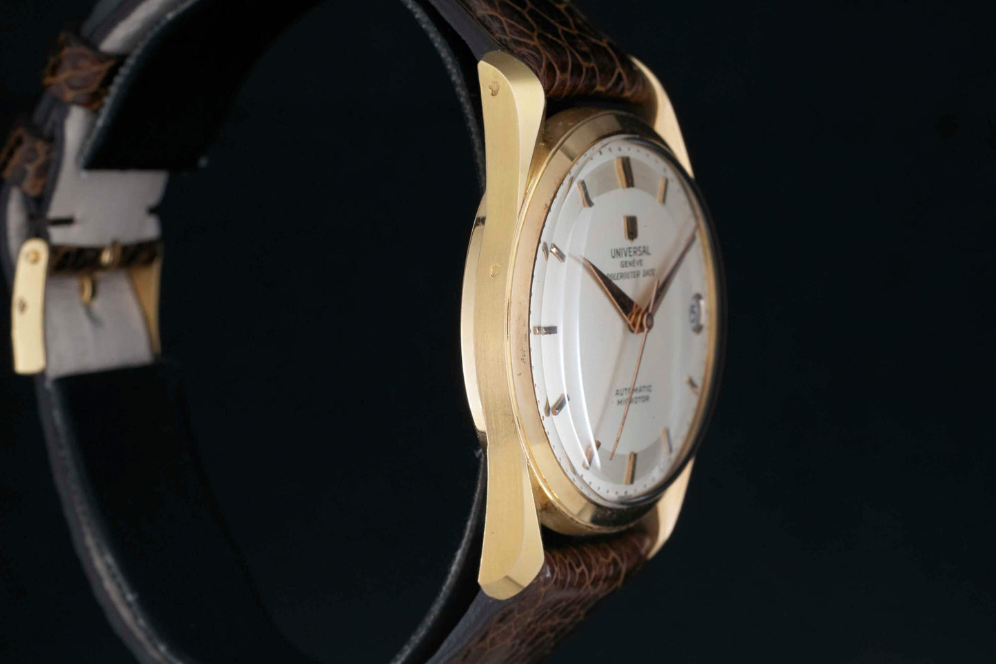 A close-up of the Universal Genève Polerouter Date dress watch by Universal Geneve, featuring a gold case, white dial, gold hour markers, and a black leather strap against a dark background.