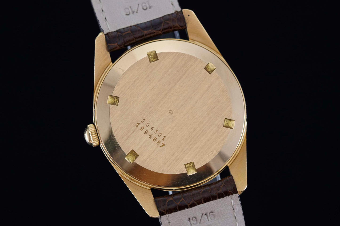 Universal Genève Polerouter Date by Universal Geneve features a gold case back with six small square markers, engraved numbers, and a brown leather strap with light gray lining, shown on a black background—a refined nod to vintage watches.