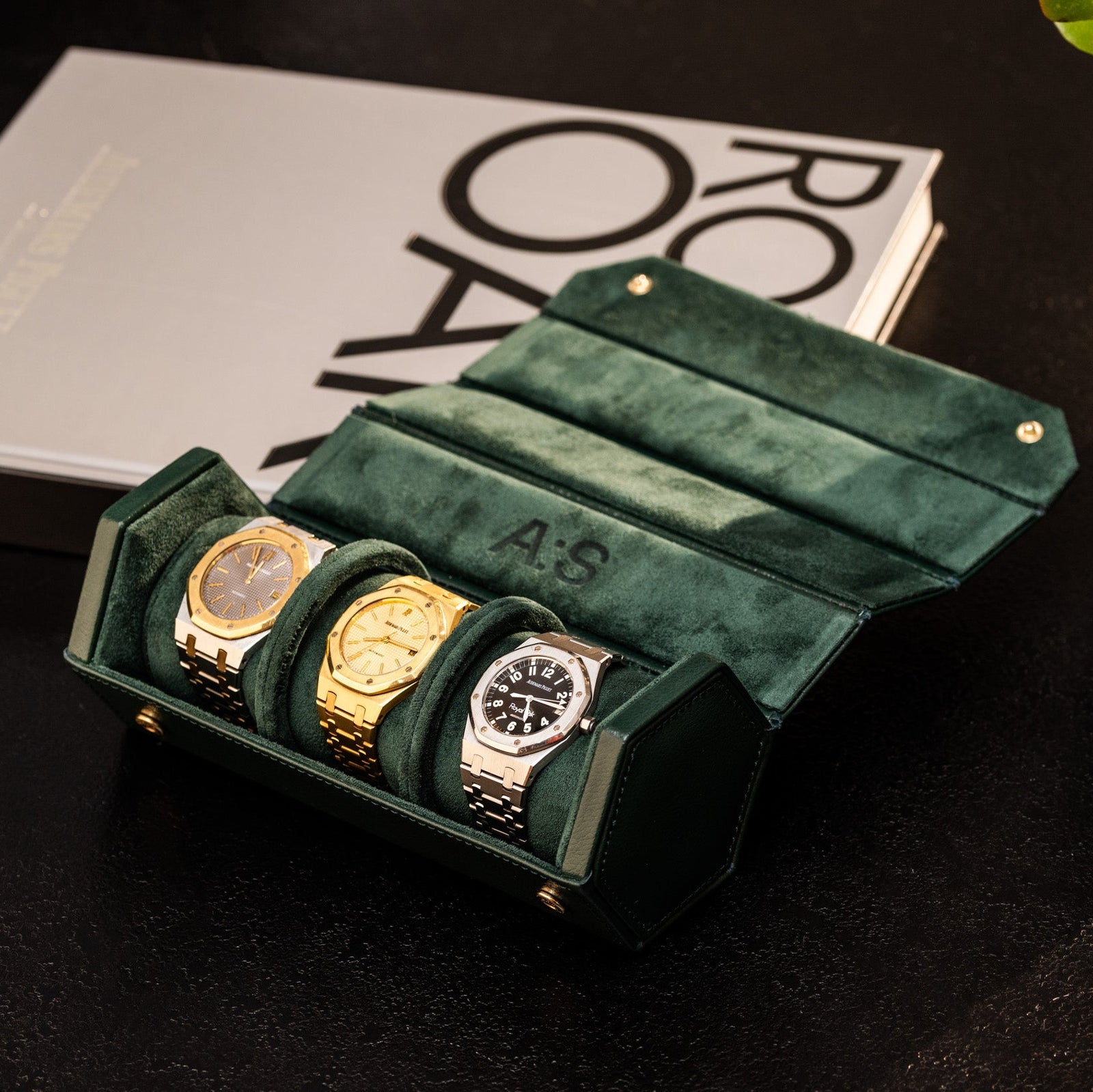A green suede WOLF Vintage Collection 3-Piece Hex Travel Tube, holding two gold and one silver watch, rests on a dark surface beside a potted succulent and a closed gray book.